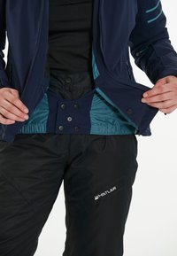 Marinblå jacka med teal-fodring öppen för att avslöja svarta byxor med flera knappar och en logo. Slät tyg med en lätt glans.