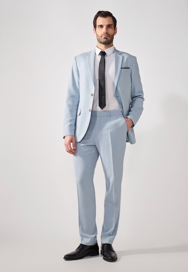 Traje azul claro con un corte estructurado, chaqueta de un solo botón, solapas de muesca y corbata negra. Combinado con zapatos de cuero negro.