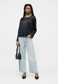 Γυναίκα με πλεγμένα μαλλιά που φοράει μαύρο σατέν πουκάμισο, ανοιχτό μπλε παντελόνι με φαρδιά πόδια, μαύρες γόβες και κρατάει μια μαύρη τσάντα.