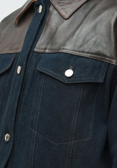 Σκούρο τζιν πουκάμισο με καφέ δερμάτινη λαιμόκοψη, διαθέτοντας ασημένια κουμπιά με σούστα και σχέδιο διπλής τσέπης. Η υφή του υφάσματος αντιπαραβάλλεται με το λείο δέρμα.