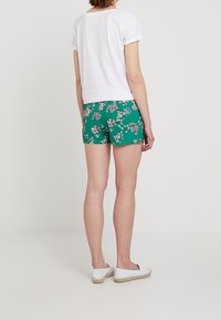 Camicia bianca a maniche corte abbinata a pantaloni corti verde con fantasia floreale, caratterizzata da un design aderente, una texture liscia e scarpe bianche piatte.