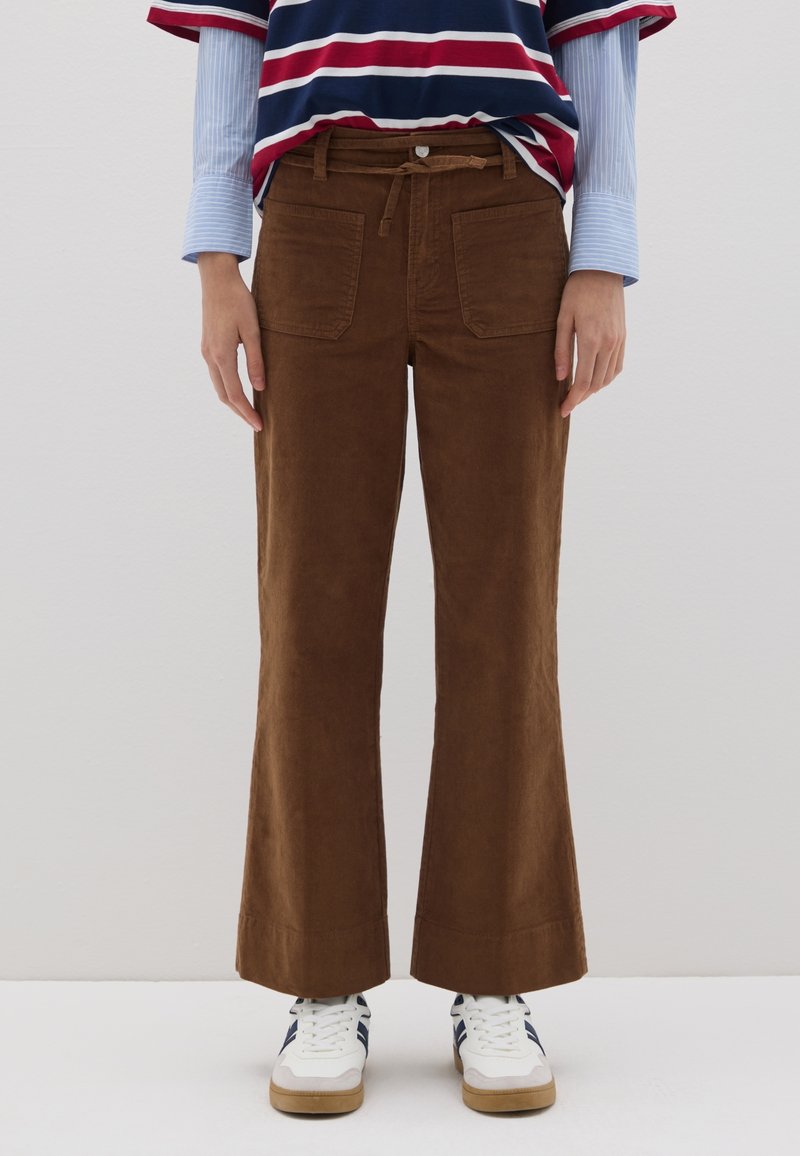 Pantalones acampanados de pana marrón con una textura suave, que cuentan con bolsillos frontales y un cordón, combinados con una camiseta a rayas y zapatillas blancas.
