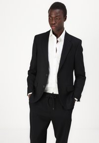 Black suit jacket with two buttons, paired with a white shirt and black drawstring trousers. Smooth fabric, tailored fit, and minimalist design.