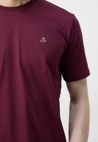 Un homme portant un t-shirt à manches courtes, rouge foncé, avec de petites lettres blanches "M O P" sur la poitrine gauche, sur un fond uni.