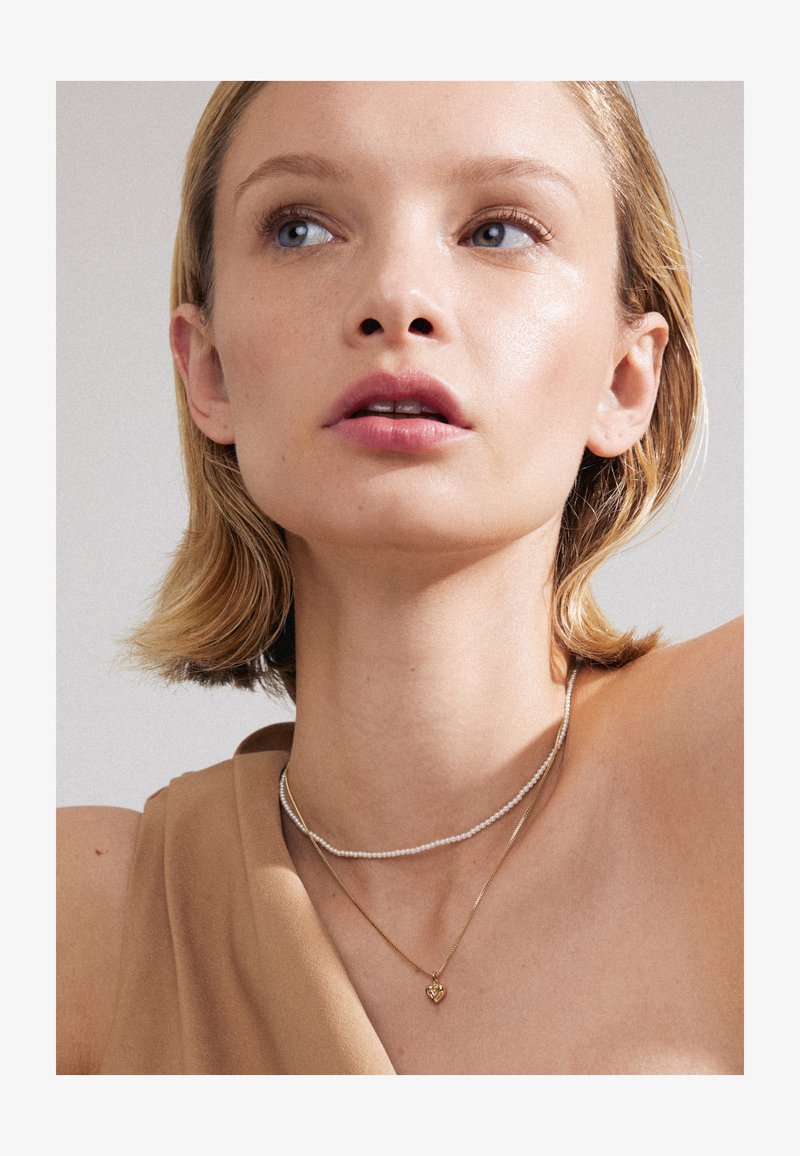 Gold heart pendant necklace layered with a delicate silver beaded choker, worn with a beige top. Skin is smooth and lightly illuminated.