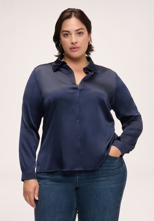 Kvinde med kort mørkt hår iført en marineblå satin-knap-op-skjorte og blå jeans, stående med den ene hånd i lommen og neutral baggrund.