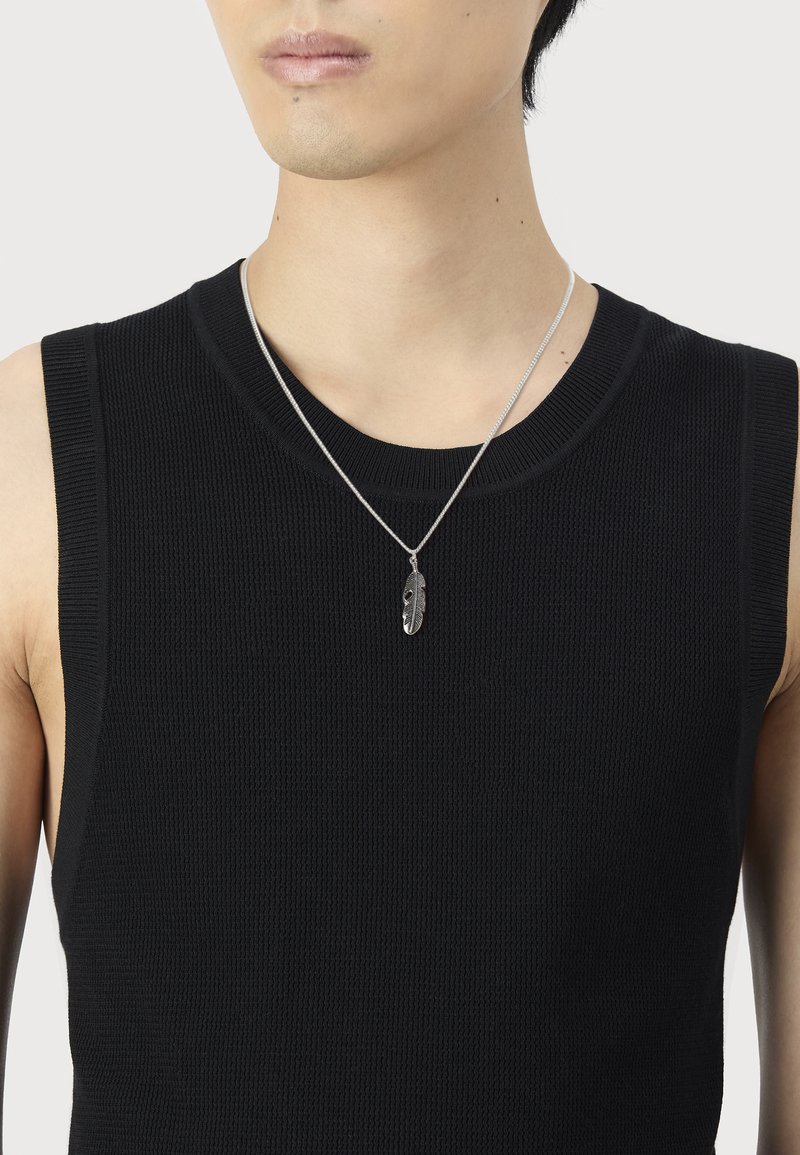 Homme portant un haut sans manches en tricot noir et un collier pendentif en argent en forme de plume sur fond blanc.