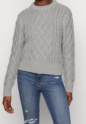 Personne portant un pull en maille torsadée gris clair et un jean bleu délavé sur un fond blanc uni.