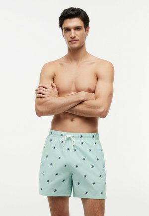 Shirtless man with crossed arms wearing light green swim shorts with small dark patterns and a white drawstring.