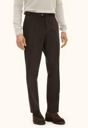 Mann trägt dunkelbraune, maßgeschneiderte Hose, hellgrauen Langarmpullover und dunkelbraune elegante Schuhe und steht vor schlichtem Hintergrund.