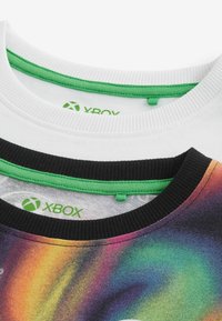 Tre T-shirt di diversi colori: una bianca, una nera con bordi verdi e un'altra con un motivo multicolore. Tutte presentano il logo di Xbox sul collo.