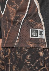 Čierny a hnedý vzorovaný atletický dres vyrobený z sieťoviny, s bielou nášivkou s logom "NPD" a "No Problemo." Viditeľn�é detaily šitia.