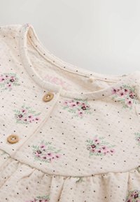 Cream baby dress with small pink floral patterns, wooden buttons near the round neckline, and subtle brown polka dots on textured fabric.