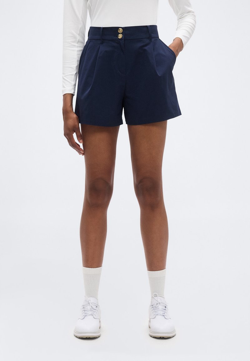 Άτομο που φοράει μπλε ναυτικό σορτς με δύο κουμπιά στη μέση, λευκό μακρυμάνικο μπλουζάκι, λευκές κάλτσες και λευκά δερμάτινα παπούτσια με κορδόνια, στέκεται με το ένα χέρι στην τσέπη.