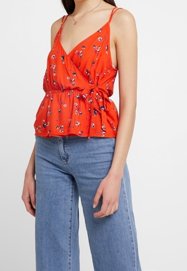 Rode bloemenprint cami-top met verstelbare bandjes, V-hals en een gesnoerd taille. Draagt bij hoge taille lichtblauwe spijkerbroek.