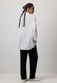 Witte blouse met lange mouwen en een losse pasvorm, gecombineerd met zwarte broek. Voorzien van een zwart haarribband met witte stippen en witte schoenen.