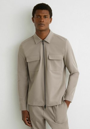 Muž v béžovej bunde na zips s klopovými vreckami a zladenými béžovými nohavicami, stojaci pred bielou textúrovanou stenou s jednou rukou vrecku.