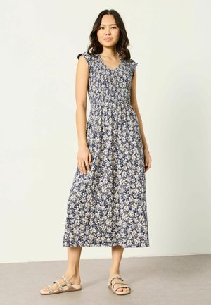 Naine seisab kandes tumelilla lillemustriga varrukateta midi kleiti ja beežeid madalaid sandaale heleda halli põranda ning valge seina taustal.