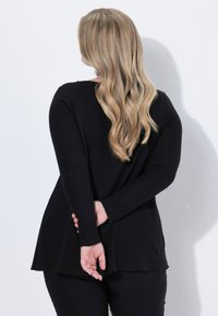 Schwarzes Langarmoberteil mit lässiger Passform und abgerundetem Saum, kombiniert mit schwarzen Leggings. Glattes Material mit minimaler Struktur, keine sichtbaren Muster.