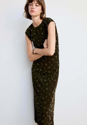 Joven mujer con el pelo corto que lleva un vestido de color verde oscuro con estampado floral, de pie con los brazos cruzados frente a un fondo blanco liso.
