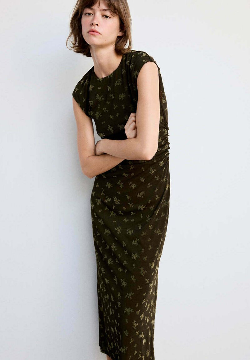 Jeune femme aux cheveux courts portant une robe vert foncé à motif floral, debout les bras croisés devant un fond blanc uni.