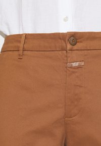 Brązowe bawełniane spodnie z zapięciem na guzik, przeszyciami i naszywką na pasku. Gładka faktura, zwężany krój.