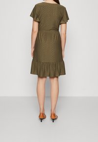 Oliivinvihreä mekko, jossa on lyhyet hihat, koristeellinen kuvioitu kangas, vyötärö vedetty sisään ja kerroksittainen röyhelöhelma. Yhdistetty ruskeisiin kenkiin.