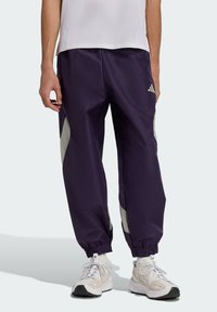 Fialové športové nohavice s elastick�ými manžetami, s striebristými bočnými panelmi a logom Adidas. Vyrobené z ľahkej, hladkej látky.