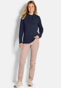 Donna sorridente con lunghi capelli biondi, vestita con una felpa blu navy, pantaloni beige e sneaker beige, in piedi con una mano in tasca.