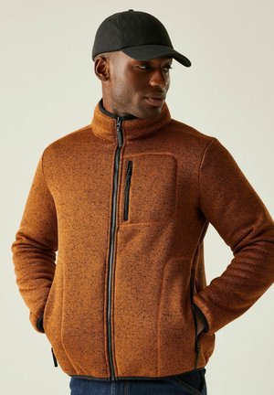 Mężczyzna w czarnej czapce i pomarańczowej kurtce na zamek z rękami w kieszeniach, patrzący w prawo na jednolitym tle.