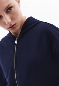 Felpa con zip di colore blu navy in morbido tessuto, con zip frontale e cappuccio, caratterizzata da una texture liscia e un design minimalista privo di motivi.