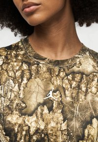 Κοντινό πλάνο ενός ατόμου που φοράει πουκάμισο με καμουφλάζ μοτίβο και λευκό λογότυπο Air Jordan στο στήθος, εστιάζοντας στον λαιμό και το κάτω μέρος του προσώπου.