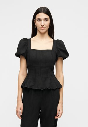 Kvinne med rett mørkt hår, iført gullringer i ørene og en svart topp med puffede ermer og bølget peplumkant, står foran en ensfarget bakgrunn.