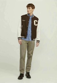 Jaqueta varsity masculina, corpo castanho com mangas brancas, apresentando um logo "C", punhos e gola listrados. Combinada com uma camisa azul e calças cinzas.
