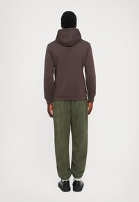 Mann som står med ryggen til iført en mørkebrun hettegenser med hetta opp og løse olivengrønne joggebukser mønstret med tekst.