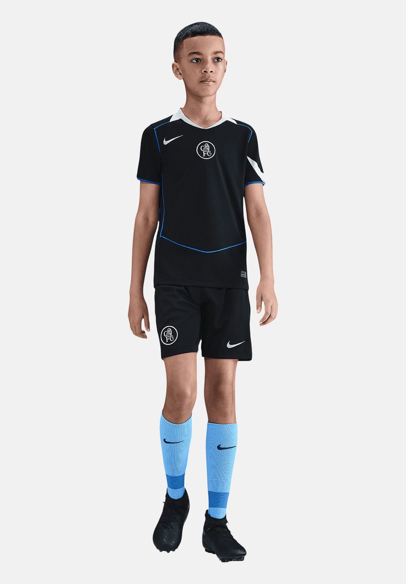 Garçon portant un maillot de football noir avec des bordures bleues, des chaussettes bleu clair et des crampons noirs, debout devant un fond uni.