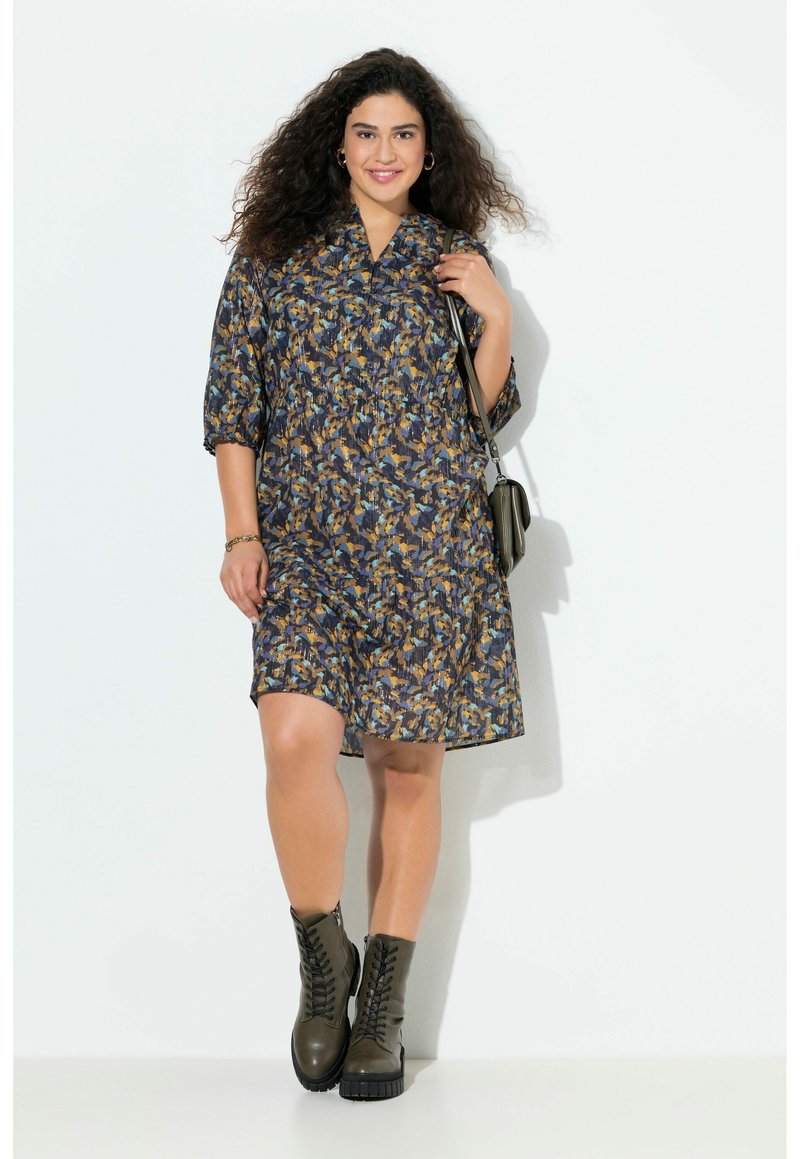 Ein Kleid mit blauen und gelben Mustern, das einen kragenförmigen Ausschnitt und ¾-Ärmel aufweist, kombiniert mit dunkelgrünen Schnürstiefeln.