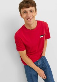 Rode katoenen T-shirt met een ronde hals, korte mouwen en een klein zwart, wit en rood logo-opdruk op de borst. Gecombineerd met blauwe jeans.