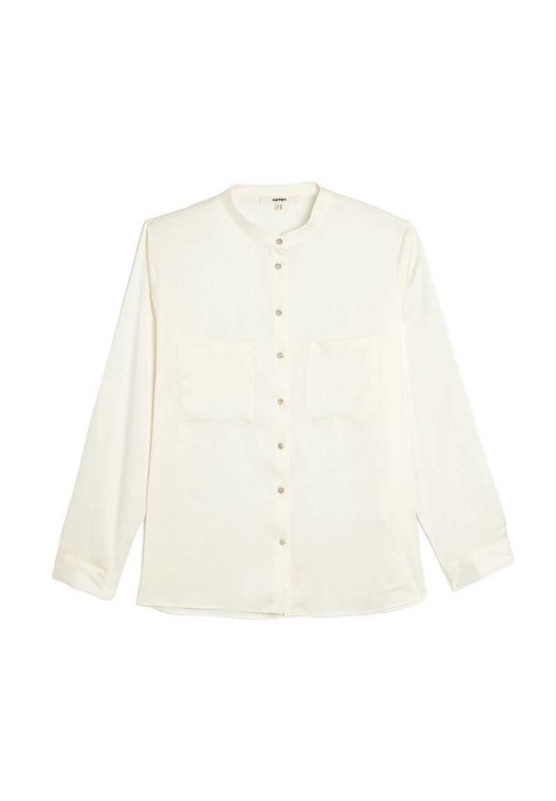 Camicia bianca a maniche lunghe con colletto rotondo, chiusura frontale con bottoni e due tasche sul petto. Tessuto morbido con una consistenza liscia.