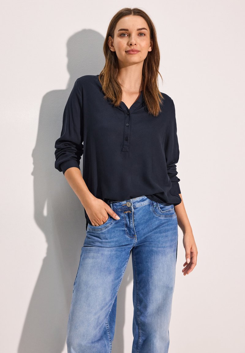 Blusa azzurra a maniche lunghe con bottoni realizzata in tessuto morbido, abbinata a jeans in denim blu chiaro con vestibilità rilassata e tasche frontali.