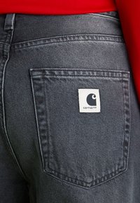Jeans de mezclilla oscura con un bolsillo trasero que lleva una etiqueta blanca con el logo de Carhartt. La tela tiene una textura ligeramente deslavada y detalles de costura limpios.