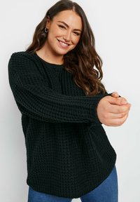 Svart stickad tröja med ett strukturerat mönster, med långa ärmar och rundad nederkant. Buren över blå jeans.