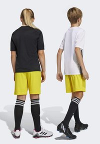 Twee kinderen in sportkleding: de één in een zwart T-shirt en gele shorts, de ander in een wit T-shirt en gele shorts. Beide dragen zwarte sokken en voetbalschoenen.