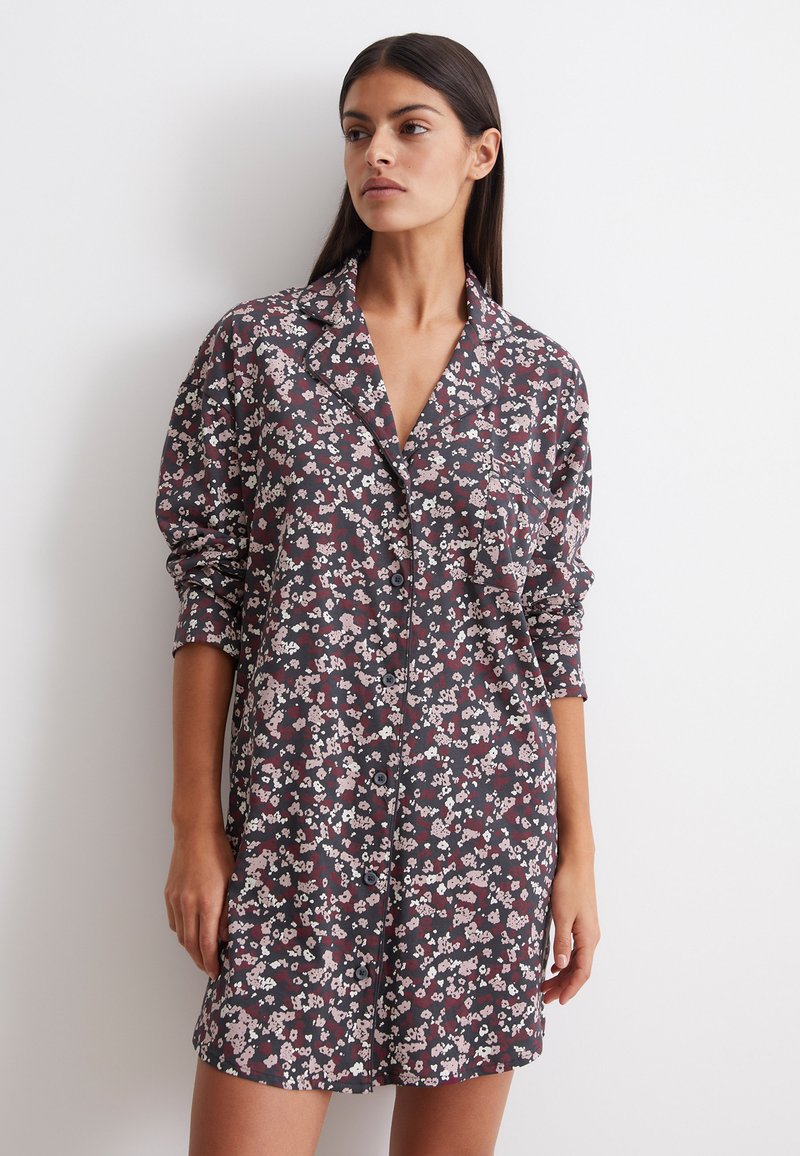 Vestito camicia floreale con uno sfondo scuro e fiori rosa e bianchi, chiusura frontale con bottoni, colletto e maniche lunghe.