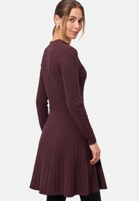 Robe en tricot bordeaux avec un motif à torsades sur le corsage, une taille cintrée et une jupe plissée évasée. Des manches longues complètent le design.