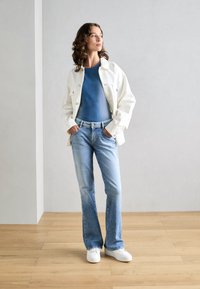 Weiße Denim-Jacke über einem blauen gerippten Top, kombiniert mit hellblauen Jeans und weißen Sneakers. Das Model steht mit den Händen in den Taschen und blickt nach links.