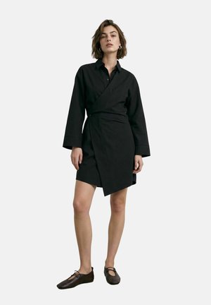 Femme portant une robe portefeuille noire à manches longues avec un ourlet asymétrique et des chaussures plates marron à lacets, debout devant un fond uni.