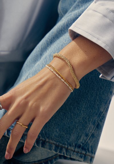 Handgelenk mit drei Armbändern: ein goldfarbenes Perlenarmband, ein dünner goldener Ring und ein perlenbesetztes Armband, getragen mit einem goldenen gedrehten Ring und Jeans.