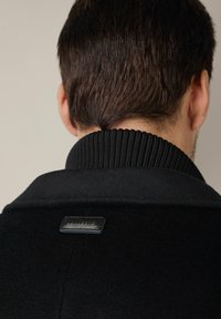 Czarny płaszcz z wyraźnym kołnierzem i żebrowanym detalem na szyi. Posiada skórzaną metkę z tyłu, gładką, wełnianą fakturę.