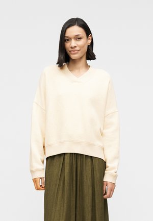 Felpa beige chiaro oversize con scollo a V, spalle scese e polsini a costine, abbinata a una gonna verde oliva texturizzata.